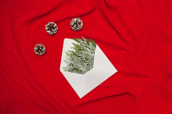 Kırmızı tuval üzerinde beyaz bir zarf içinde donmuş selvi ve çam kozalakları ile yeni yıl kartı göndermeye hazır bir Noel kompozisyonu. Asgari Noel fikri.