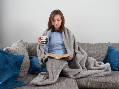 Evdeki kanepede oturup kitap okuyan yumuşak bir battaniyeye sarılmış güzel bir genç kadın. Elinde bir fincan sıcak çay tutuyor..