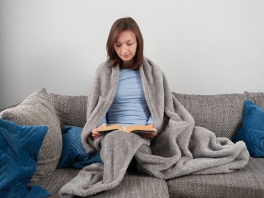Yumuşak bir battaniyeye sarılı güzel genç bir kadın evdeki kanepede oturuyor ve kitap okuyor..