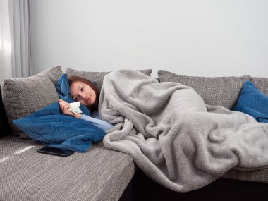 Yumuşak bir battaniyeye sarılı hasta, genç, soğuk bir kadın evdeki kanepede yatıyor. Kız elinde bir mendil tutuyor..