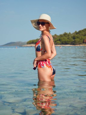 Renkli mayolu ve güneş şapkalı genç, çekici bir kız turkuaz suyla denizde duruyor. Sahilde güzel ve zarif bir kadın.
