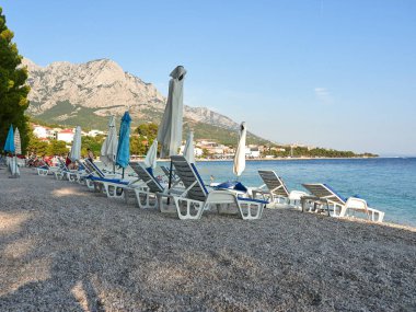 Baska Voda, Dalmaçya, Hırvatistan - 22 Temmuz 2021: Hırvat kentinde turistlerle birlikte İkovac plajı. Adriyatik Denizi 'nde güneş şemsiyeleri ve plaj şezlongları.