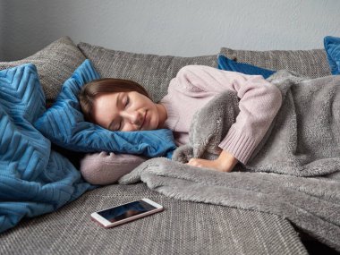 Genç bayan oturma odasındaki kanepede şekerleme yapıyor. Gri bir battaniyeye sarılı bir kız, mavi bir yastığa sarılıp kanepede uyuyor. Önünde bir akıllı telefon var..