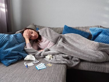Genç hasta bir kız battaniyenin altında kanepede yatıyor. Başı ağrıyan bir kadın başını tutuyor. Termometre, tablet paketleri ve mendiller kanepede yatıyor.