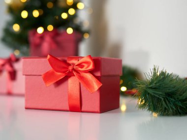 Kırmızı hediye kutusunu kapat. Kırmızı kurdeleli ve fiyonklu güzel bir hediye kutusu. Yeşil Noel ağacı ve ışıkları olan bir arka plan. Noel zamanı ve hediye zamanı
