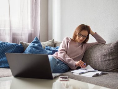 Gözlüklü genç bir kadın kanepede oturmuş açık bir kitap düşünüyor. Kız oturma odasında oturuyordu. Kafasını eliyle destekliyor ve düşünüyor. Masada bir dizüstü bilgisayar var..