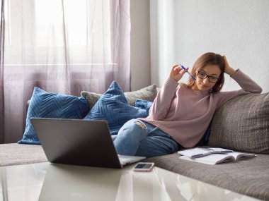 Gözlüklü genç bir kız öğrenci kanepeye oturur ve açık bir kitap üzerinde düşünür. Kız evde ders çalışıyor..