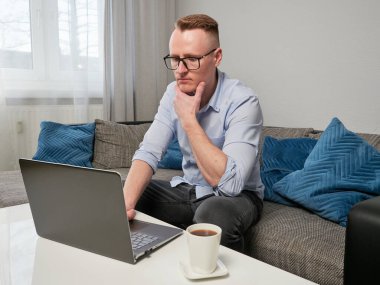 Gözlüklü genç, düşünceli bir adam kahve masasındaki kanepede oturuyor. Dizüstü bilgisayar kullanıyor. Onun yanında bir fincan kahve var. Merkez Ofis.