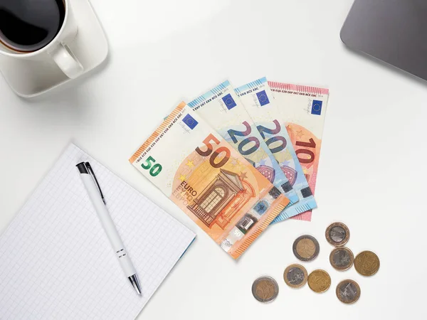 Masada duran Euro banknotlarının ve paraların en üst görüntüsü. Yanında bir fincan kahve, içinde kalem ve dizüstü bilgisayar olan bir defter var. Beyaz arkaplan.