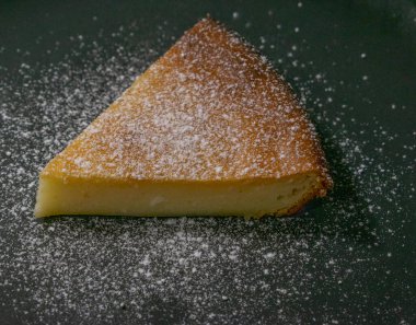 Siyah arka planda bir parça peynirli kek üzerine pudra şekeri serpiştirilmiş.