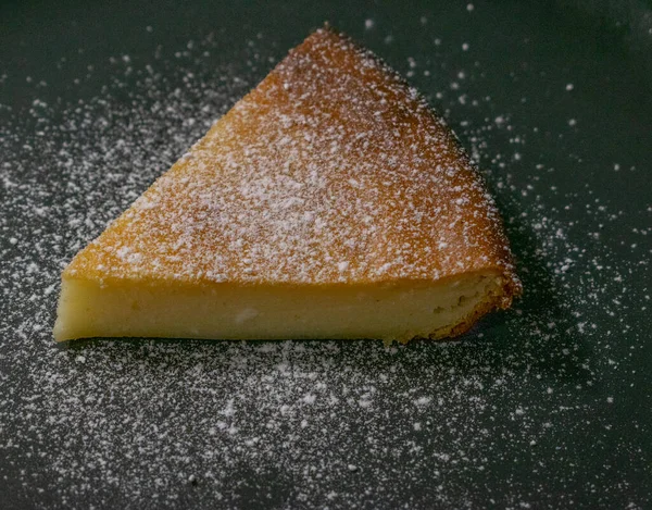 Siyah arka planda bir parça peynirli kek üzerine pudra şekeri serpiştirilmiş.