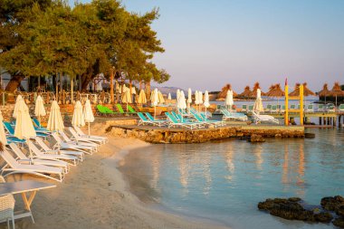 Turkuaz deniz suyu, güneş pansiyonları ve şemsiyelerle beyaz kumlu sahilde gün doğumu.