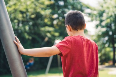 Kırmızı tişörtlü bir çocuk metal bir direğe yaslanmış oyun parkındaki teleferiğe binen bir çocuğu izliyor.