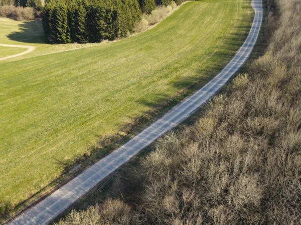 Top view of a winding road between a grass field and trees without leaves in spring