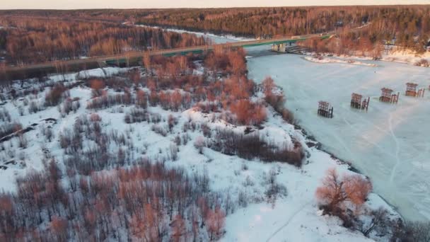 Прекрасний зимовий пейзаж з замерзлою річкою і мостом на заході сонця, вид з повітря . — стокове відео