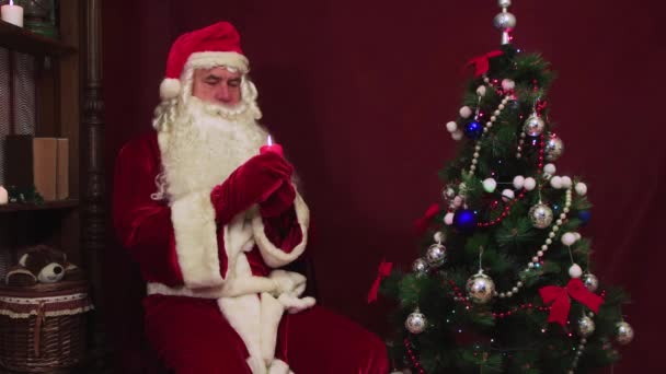 Papai Noel moderno segura uma vela vermelha acesa em sua mão e parabeniza. — Vídeo de Stock