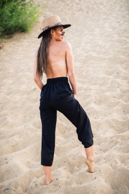 Siyah pantolonlu ve şapkalı çıplak esmer model, kameranın önünde kumda poz veriyor. Yakın çekim ve uzun menzilli çekim.