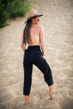 Siyah pantolonlu ve şapkalı çıplak esmer model, kameranın önünde kumda poz veriyor. Yakın çekim ve uzun menzilli çekim.