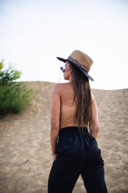 Siyah pantolonlu ve şapkalı çıplak esmer model, kameranın önünde kumda poz veriyor. Yakın çekim ve uzun menzilli çekim.