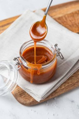 Ahşap tahtada cam kavanozda ev yapımı tuzlu karamel sosu, gri arka plan. Karamel sosu elinde kaşıktan damlıyor..