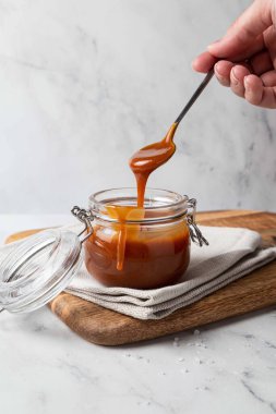 Ahşap tahtada cam kavanozda ev yapımı tuzlu karamel sosu, gri arka plan. Karamel sosu elinde kaşıktan damlıyor..