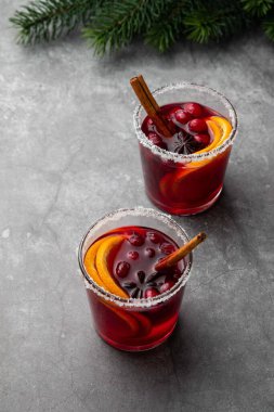 Kış için sıcak sıcak şarap, kızılcık, baharat, tarçın ve portakal dilimleri gri masada iki bardak içinde. Noel içkisi. Noel ağacı dallarıyla şenlikli bir konsept. Seçici odak.