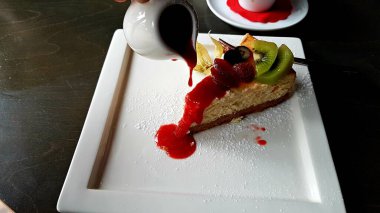 Büyük bir dilim pasta ve beyaz bir tabakta vişne reçelli sos kabı..