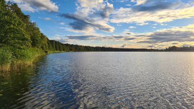 Geniş nehir üzerinde bulutlu gökyüzünde gün batımı ve kıyılarında yeşil orman.