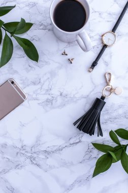 Beyaz tezgahın üstünde sade kahve olan beyaz bir kupa. Yakınlarda beyaz kremalar var. Bir telefon, siyah bir şapka ve siyah kadın aksesuarları. Üst görünüm.