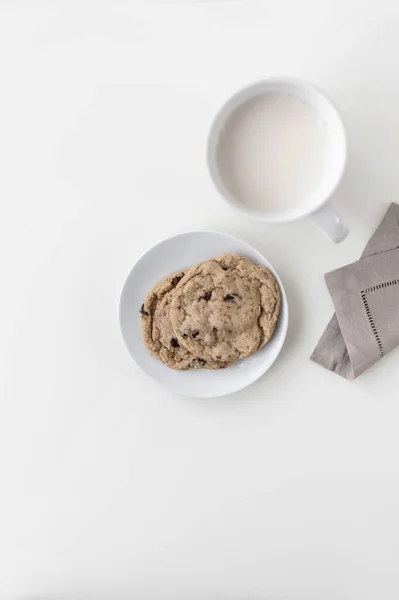 A white mug with milk on a white countertop. There is a napkin next to it and one cookie on a plate. Top view — Stock Photo, Image