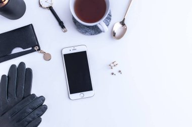 Beyaz tezgahın üstünde sade kahve olan beyaz bir kupa. Yakınlarda beyaz kremalar var. Bir telefon, siyah bir şapka ve siyah kadın aksesuarları. Üst görünüm.