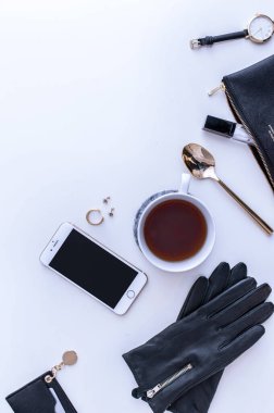 Beyaz tezgahın üstünde sade kahve olan beyaz bir kupa. Yakınlarda beyaz kremalar var. Bir telefon, siyah bir şapka ve siyah kadın aksesuarları. Üst görünüm.