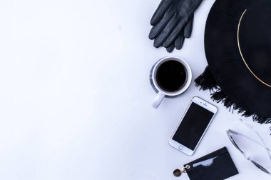 Beyaz tezgahın üstünde sade kahve olan beyaz bir kupa. Yakınlarda beyaz kremalar var. Bir telefon, siyah bir şapka ve siyah kadın aksesuarları. Üst görünüm.