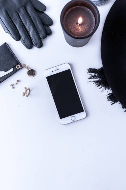 Beyaz tezgahın üstünde sade kahve olan beyaz bir kupa. Yakınlarda beyaz kremalar var. Bir telefon, siyah bir şapka ve siyah kadın aksesuarları. Üst görünüm.