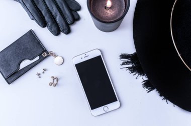 Beyaz tezgahın üstünde sade kahve olan beyaz bir kupa. Yakınlarda beyaz kremalar var. Bir telefon, siyah bir şapka ve siyah kadın aksesuarları. Üst görünüm.