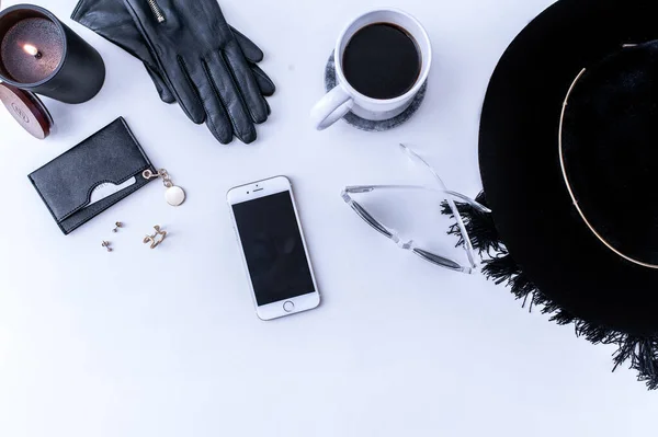 A white mug with black coffee on a white countertop. There are white meringues nearby. A phone, a black hat and black womens accessories. Top view. — Stock Photo, Image