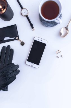 Beyaz tezgahın üstünde sade kahve olan beyaz bir kupa. Yakınlarda beyaz kremalar var. Bir telefon, siyah bir şapka ve siyah kadın aksesuarları. Üst görünüm.