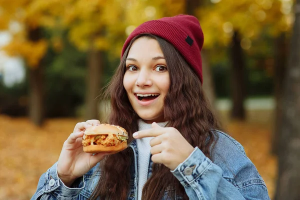 Kot ceketli ve elinde hamburgerle kırmızı şapkalı bir kız kameraya bakar ve sonbahar parkında gülümser.