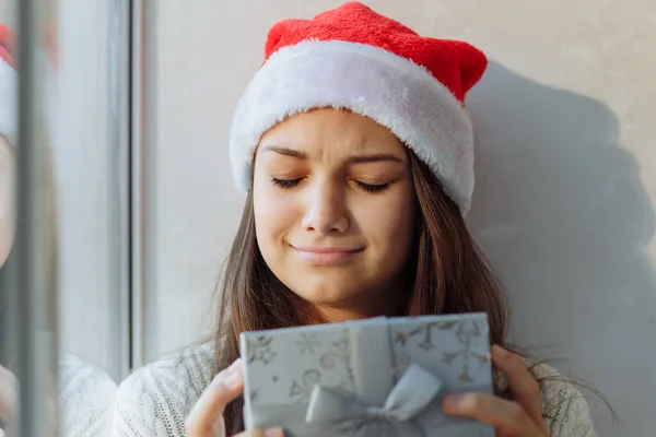 Noel şapkalı bir kız hediye kutusuyla pencerenin yanında.