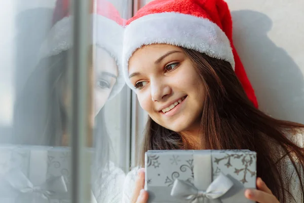 Mutlu kız Noel şapkalı, hediye kutusuyla pencerenin yanında.