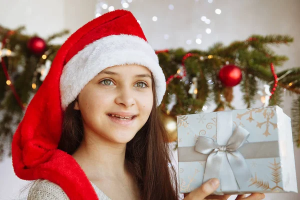 Noel şapkalı gülümseyen bir kız elinde bir hediye kutusu tutuyor.