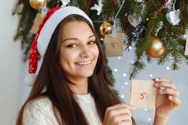 Noel şapkalı gülümseyen bir kız. Elinde bir takvim var. Noel ağacının yanında.
