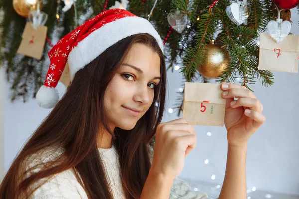 Noel şapkalı gülümseyen bir kız. Elinde bir takvim var. Noel ağacının yanında.