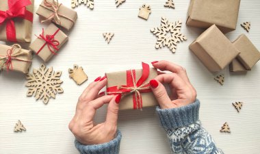 Kadının elleri Noel hediyesini kırmızı kurdele ve kenevir ipiyle el işi kağıdına sarmış. El yapımı Noel hediye kutusunda fiyonk yapıyorum. Tahta masa, bayram hediyeleri ve tahta kar taneleri. Üst görünüm.