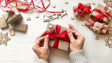 Kadının elleri Noel hediyesini kırmızı kurdeleyle el sanatları kağıdına sarmış. El yapımı Noel hediye kutusunda fiyonk yapıyorum. Tatil hediyeleri, tahta kar taneleri ve yıldızlı ahşap masa. Üst görünüm.