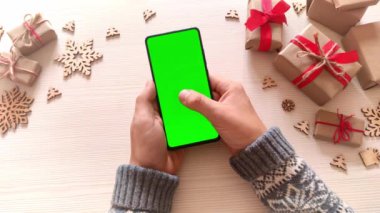 Elinde yeşil ekranlı akıllı telefon tutan ve Noel arkaplanına hediyelerle dokunan bir adam. Chroma Key 'li cep telefonu. Ellerini kapat..