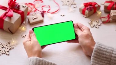 Noel arkaplanında hediyeli, yatay yeşil ekranlı akıllı telefonu olan bir kadın. Chroma Key 'li cep telefonu. Ellerini kapat..