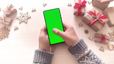Elinde dikey yeşil ekranlı akıllı telefon tutan ve Noel arkaplanına hediyelerle dokunan bir adam. Chroma Key 'li cep telefonu. Ellerini kapat..