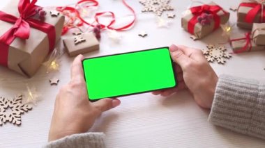 Noel arkaplanında hediyeli, yatay yeşil ekranlı akıllı telefonu olan bir kadın. Chroma Key 'li cep telefonu. Ellerini kapat..