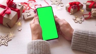 Elinde yeşil ekran olan akıllı telefonu tutan ve Noel arkaplanına hediyelerle dokunan bir kadın. Chroma Key 'li cep telefonu. Ellerini kapat..